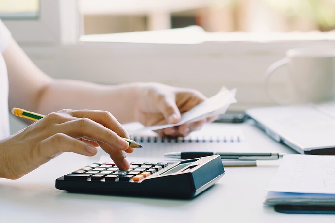 A person is using a calculator and holding receipts while working at a desk with a laptop, notebooks, and a cup of coffee. (Captioned by AI)